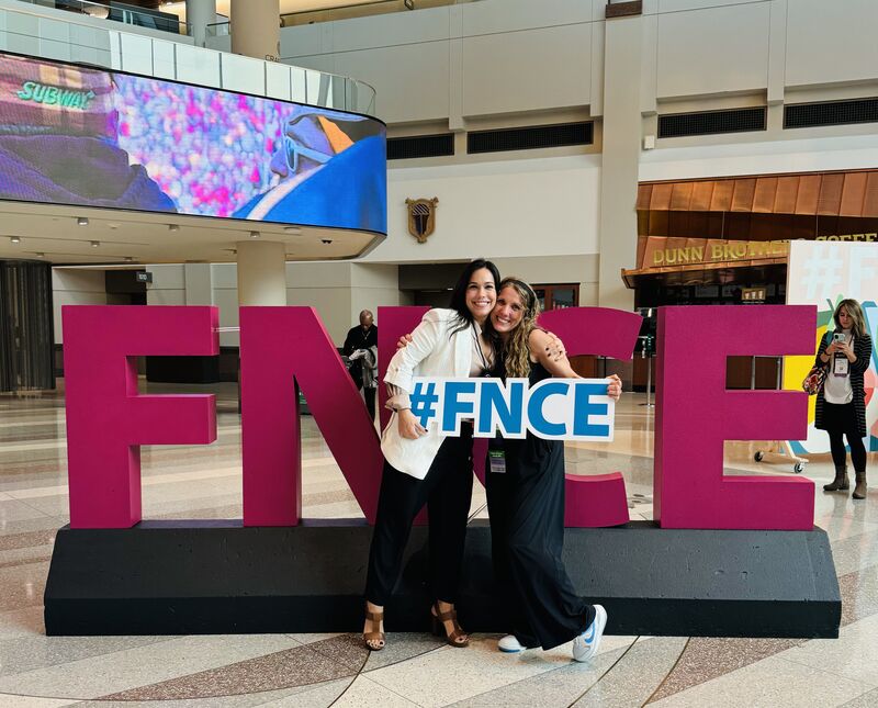 A woman with dark hair wearing a white blazer and black pants hugging a woman with blonde curly hair wearing a black jumpsuit while they both hold a white and blue sign that reads FNCE.