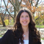 A woman with long brown hair that is curled wearing a black blazer over a white shirt with a silver heart necklace.