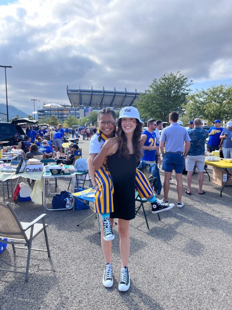 A woman with brown hair wearing a black dress and a white Pitt bucket hat holding a young girl on her back who has her hair in braids and is wearing a white t shirt and blue and gold overalls.
