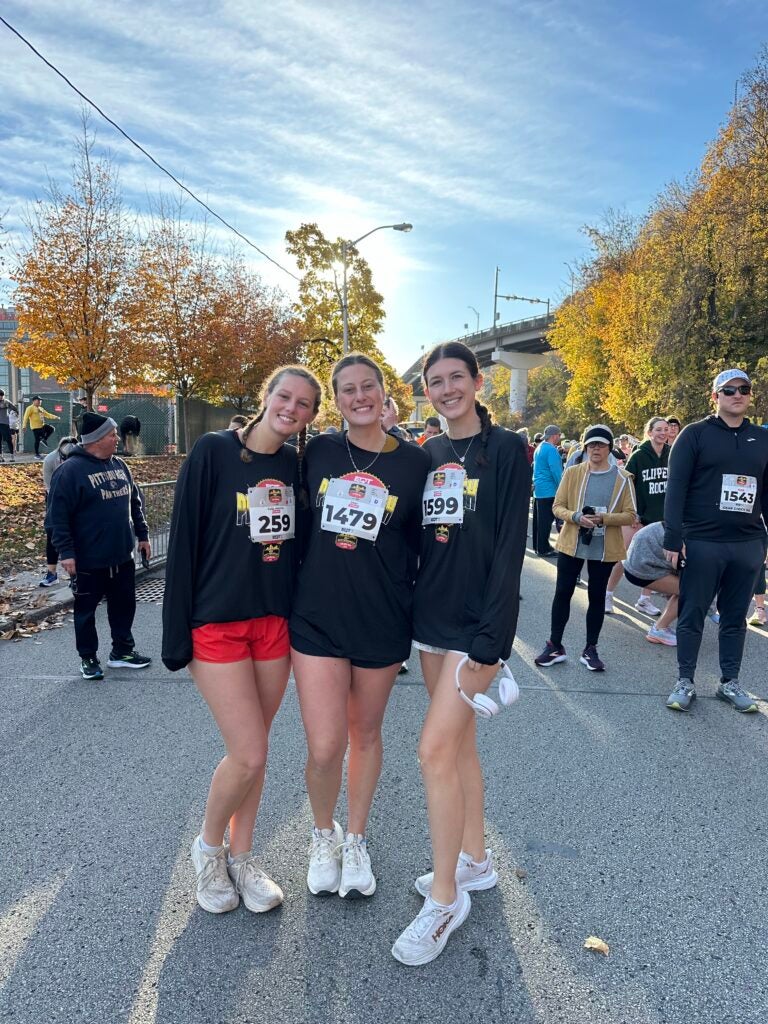 Three women with brown hair pulled back in braids wearing navy blue long sleeve shirts and shorts with white tennis shoes.