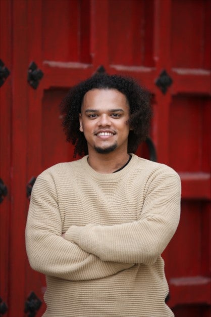 Man with dark curly hair wearing a tan sweater in front of a red door.