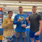 A man with short brown hair wearing a yellow short and blue and white shorts, a man with short brown hair wearing a blue shirt and gray shorts and a man with short blonde hair wearing a dark gray shirt and dark blue shorts all standing together holding cups and packages of food.