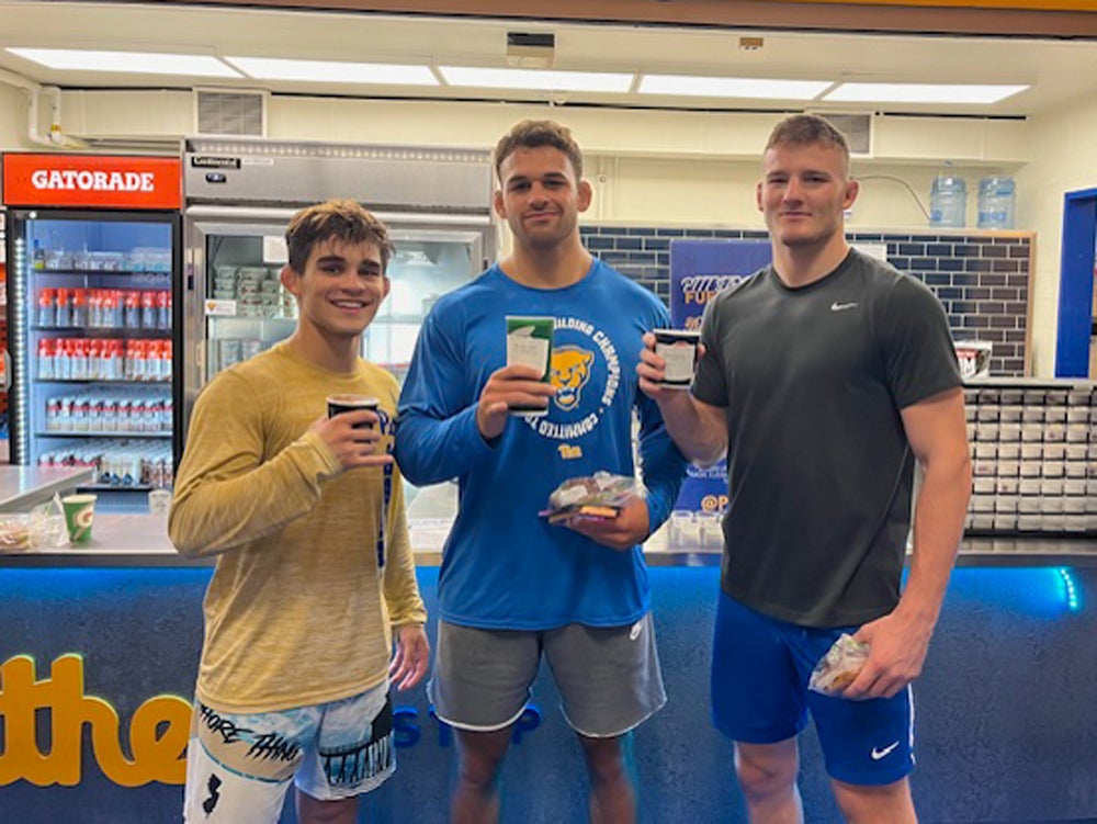 A man with short brown hair wearing a yellow short and blue and white shorts, a man with short brown hair wearing a blue shirt and gray shorts and a man with short blonde hair wearing a dark gray shirt and dark blue shorts all standing together holding cups and packages of food.
