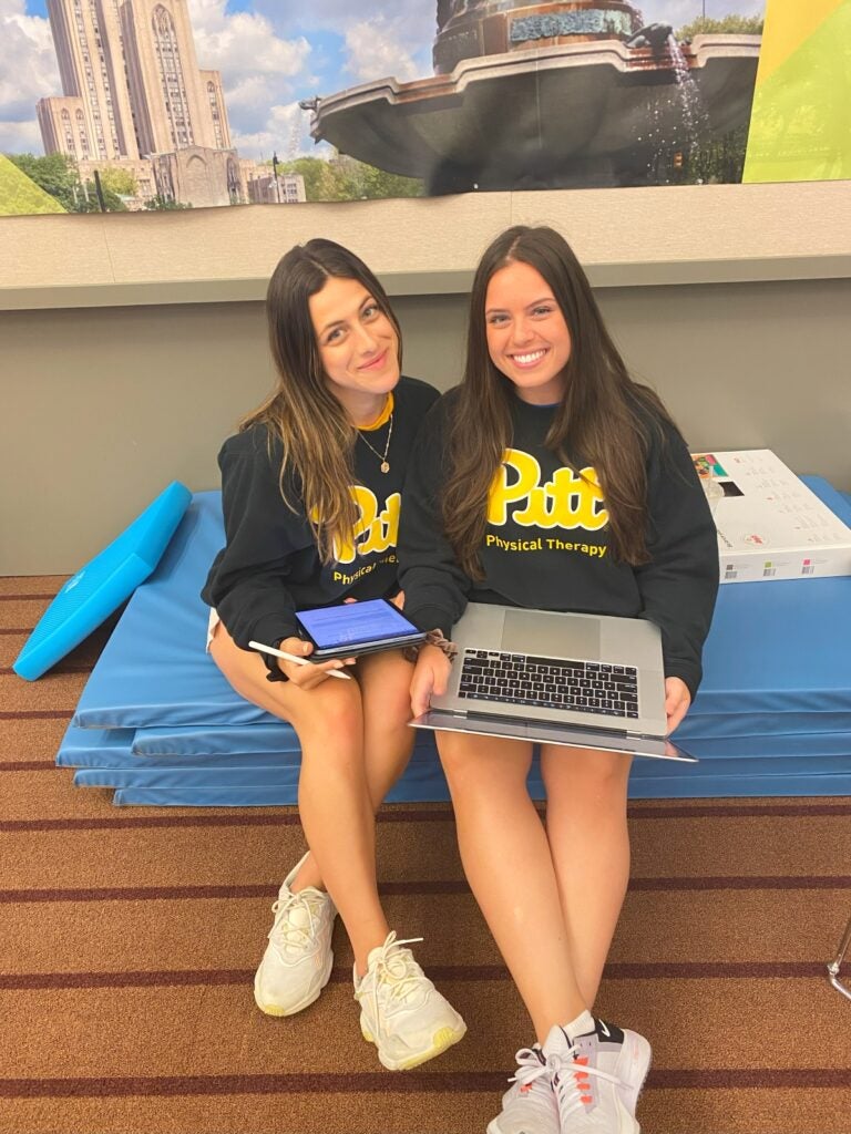 Two women with dark brown hair wearing black Pitt sweatshirts sitting together while holding laptops.