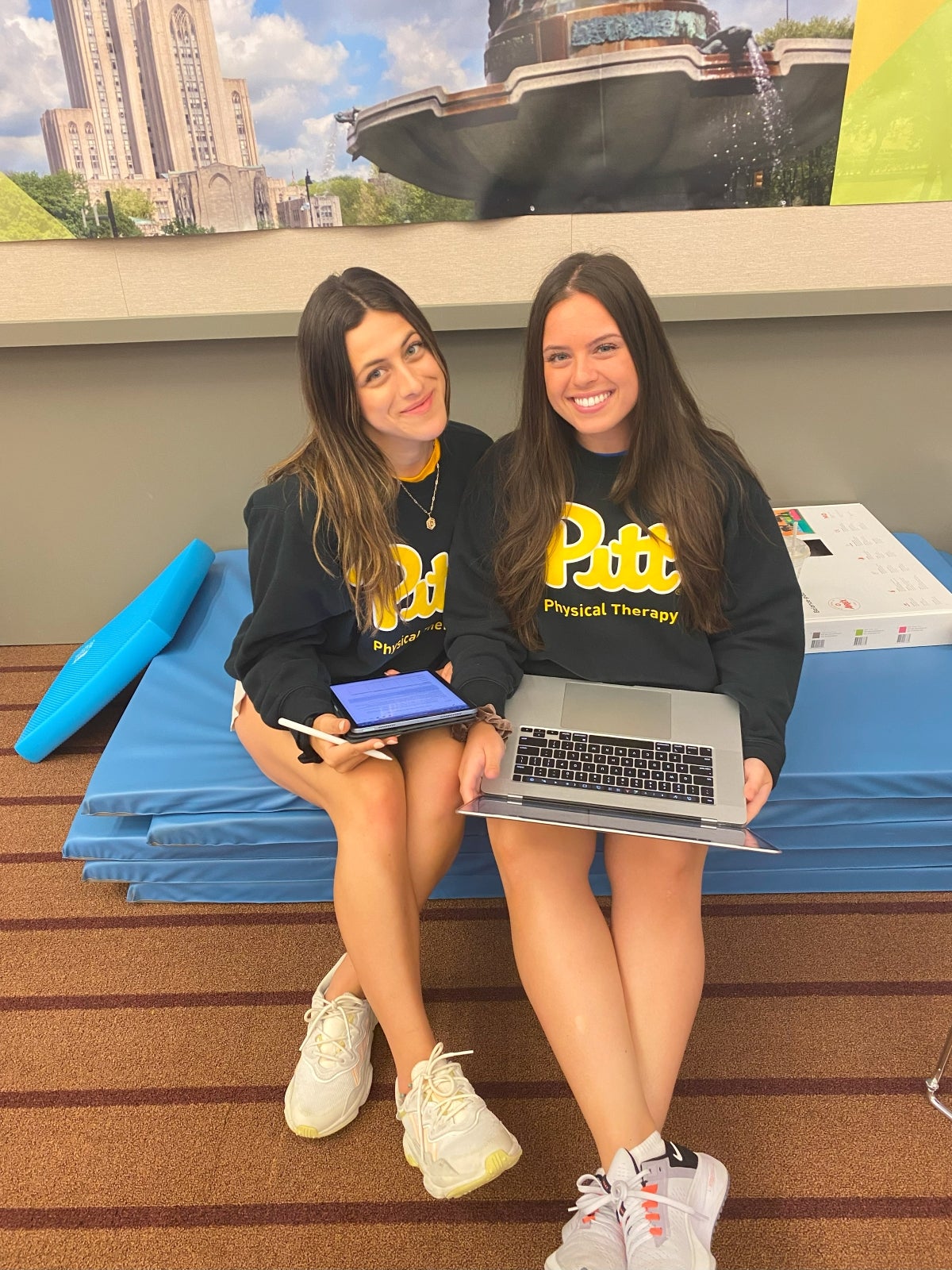 Two women with dark brown hair wearing black Pitt sweatshirts sitting together while holding laptops.