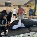 A woman with very light blonde hair wearing a black jacket over a black and white shirt and dark blue glasses stands over a woman wearing a black hijab, a black and white striped long sleeve shirt and black pants who is sitting on a stool with her hand on the head of a woman with long black braids wearing a light gray shirt and black leggings who is laying on a table.