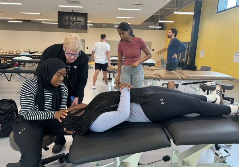 A woman with very light blonde hair wearing a black jacket over a black and white shirt and dark blue glasses stands over a woman wearing a black hijab, a black and white striped long sleeve shirt and black pants who is sitting on a stool with her hand on the head of a woman with long black braids wearing a light gray shirt and black leggings who is laying on a table.