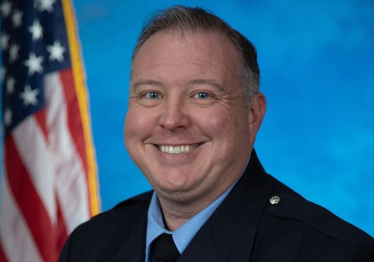 A man with short dark and gray hair wearing a black coat over a light blue collared shirt and black tie sitting in front of an American flag.