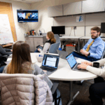 A group of women sitting around a table with computers alongside a man with brown hair, a beard and glasses wearing a blue buttoned up shirt and a yellow tie while they all look toward a tv monitor.