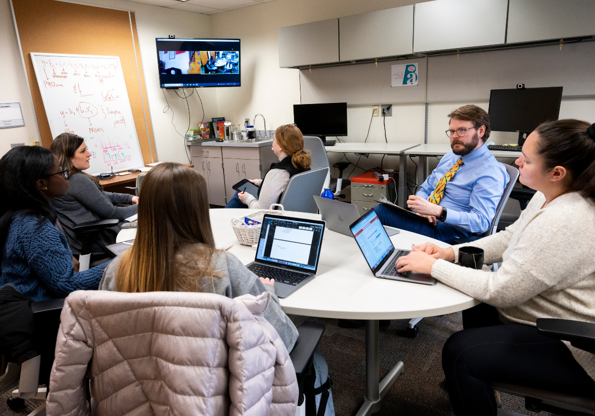 A group of women sitting around a table with computers alongside a man with brown hair, a beard and glasses wearing a blue buttoned up shirt and a yellow tie while they all look toward a tv monitor.