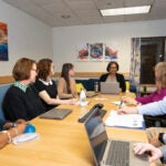 A group of men and women sitting around a long table with laptops, papers and pens while they are all looking at a woman with short black hair and black glasses who is speaking.
