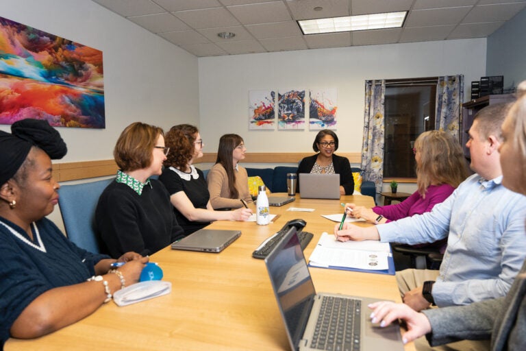 A group of men and women sitting around a long table with laptops, papers and pens while they are all looking at a woman with short black hair and black glasses who is speaking.