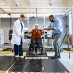 A woman with blonde hair pulled into a bun wearing a white lab coat over navy blue scrubs standing and a man with light blue collared shirt and gray jeans standing together looking at a woman in between them who has short white hair and is wearing a red shirt and a black pants while sitting in a wheelchair on a ramp.