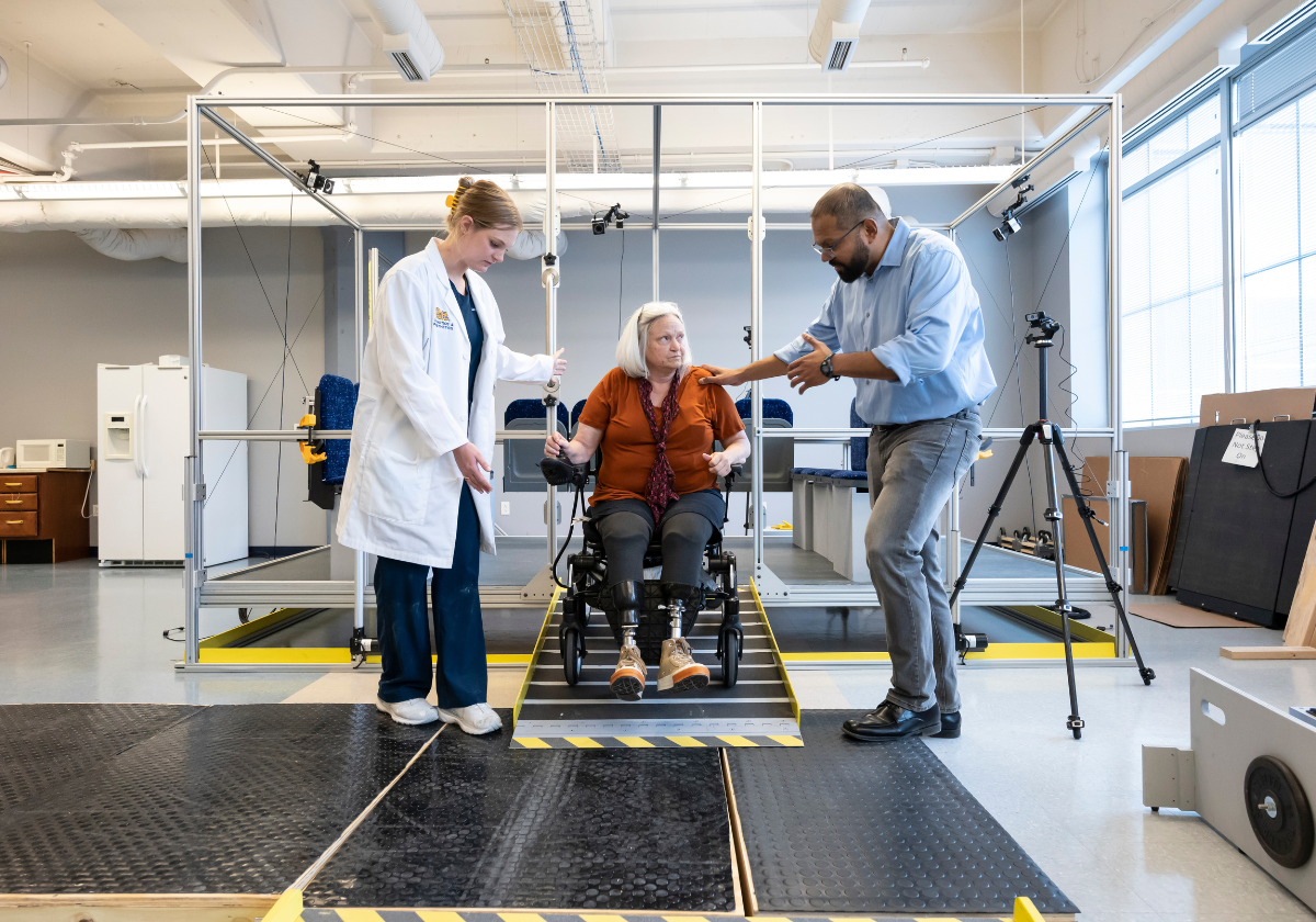 A woman with blonde hair pulled into a bun wearing a white lab coat over navy blue scrubs standing and a man with light blue collared shirt and gray jeans standing together looking at a woman in between them who has short white hair and is wearing a red shirt and a black pants while sitting in a wheelchair on a ramp.