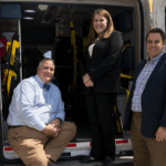 A man with short gray hair wearing a blue long sleeve shirt with a blue bow tie and tan pants, a woman with auburn hair wearing a black pantsuit and a man with short dark hair wearing a dark blue suit jacket over a plaid collared shirt and tan pants all gathered together at the back of an emergency vehicle.