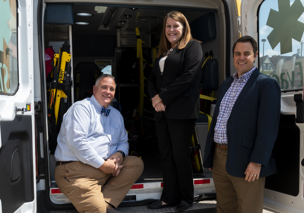 A man with short gray hair wearing a blue long sleeve shirt with a blue bow tie and tan pants, a woman with auburn hair wearing a black pantsuit and a man with short dark hair wearing a dark blue suit jacket over a plaid collared shirt and tan pants all gathered together at the back of an emergency vehicle.