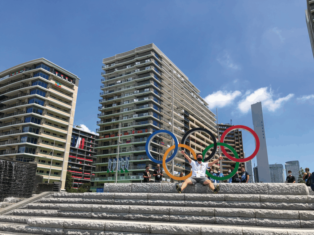 A man with short dark hair wearing a dark face mask, a white polo and gray shorts sits on cement steps in front of a sign of five different colored circles, depicting the Olympics logo.