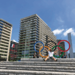 A man with short dark hair wearing a dark face mask, a white polo and gray shorts sits on cement steps in front of a sign of five different colored circles, depicting the Olympics logo.