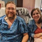 A man with auburn hair and a beard wearing clear-rimmed glasses and a blue hospital gown sitting next to a woman with short brown hair wearing a jean jacket over a red shirt.