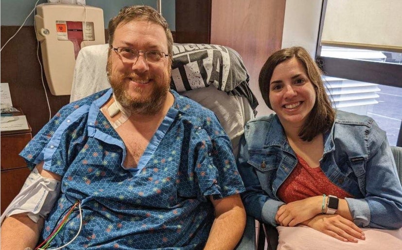A man with auburn hair and a beard wearing clear-rimmed glasses and a blue hospital gown sitting next to a woman with short brown hair wearing a jean jacket over a red shirt.