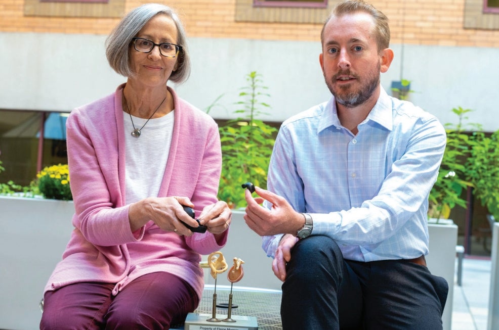 A woman with short gray hair wearing dark glasses, a white cardigan over a white top and maroon pants sitting next to a man with light brown hair and a beard wearing a light blue collared shirt and dark gray pants each holding auditory devices.