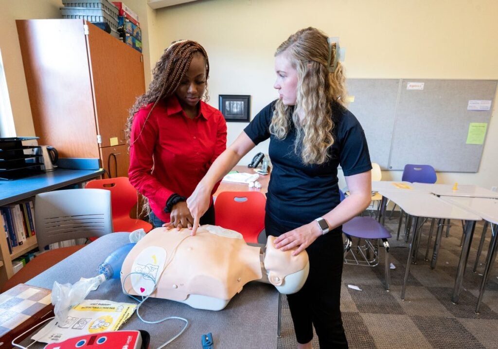 A woman with long brown braids wearing a red collared shirt and a woman with long curly blond hair wearing a black polo working together on a medical dummy.
