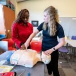 A woman with long brown braids wearing a red collared shirt and a woman with long curly blond hair wearing a black polo working together on a medical dummy.