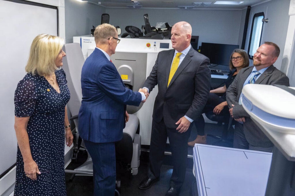 A man in a dark blue suit with short, light brown hair and glasses shakes the hand of a man with very short hair wearing a black suit and a yellow tie while two women and a man to the sides of them look on.