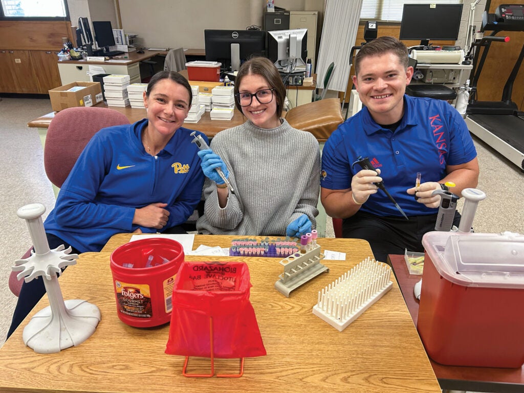 A woman with dark hair pulled back wearing a blue Pitt zip-up; a woman with brown hair pulled back wearing black glasses and a gray sweater; and a man with brown hair wearing a blue polo shirt sit together at a table while handling biospecimens.