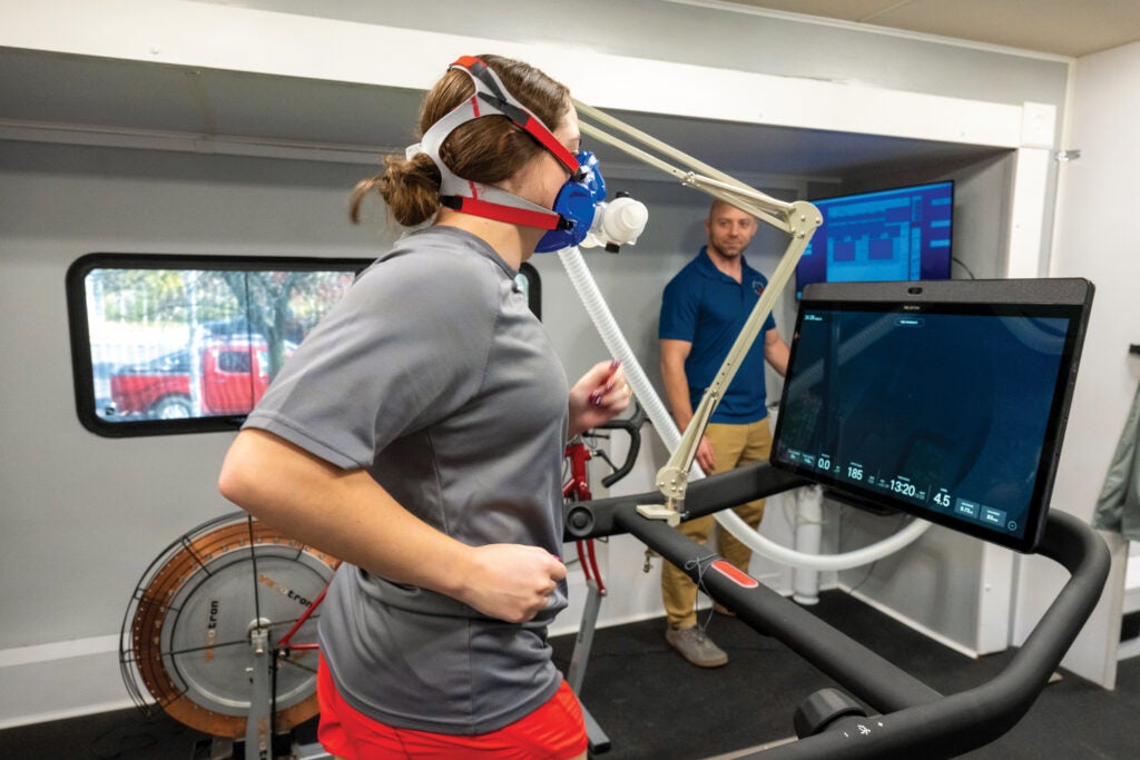 A woman wearing a gray shirt and bright red shorts wears a mask over her head that is attached to a treadmill she is on, while a man in a blue shirt and khaki pants looks on.