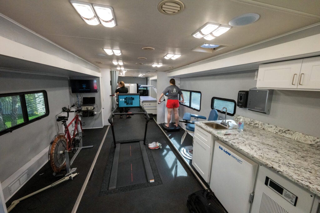 An inside of a mobile research vehicle (MRV) where exercise equipment, a sink and counter, and imaging equipment are displayed while a couple people stand in the back.