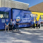 A group of men and women stand in front of a large, blue mobile research laboratory (MRV) outside.