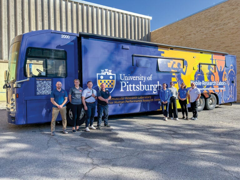 A group of men and women stand in front of a large, blue mobile research laboratory (MRV) outside.