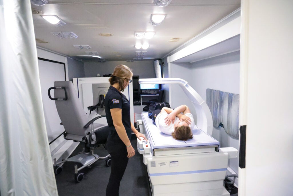 A woman with short, dark blonde hair wearing a black shirt and black pants stands at a medical imaging station where an individual is laying down.