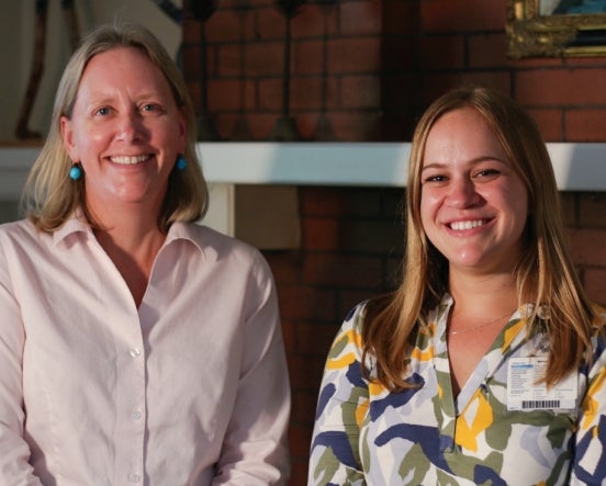A woman with short blonde hair and blue earring wearing a light pink collared shirt standing next to a woman with dark blonde hair wearing a blouse with light blue, dark blue, green and yellow designs.