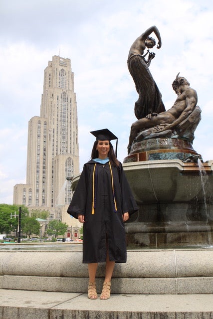 A woman with brown hair wearing a black graduation gown and cap and tan shoes standing in front of the Cathedral of Learning and a fountain with statues.