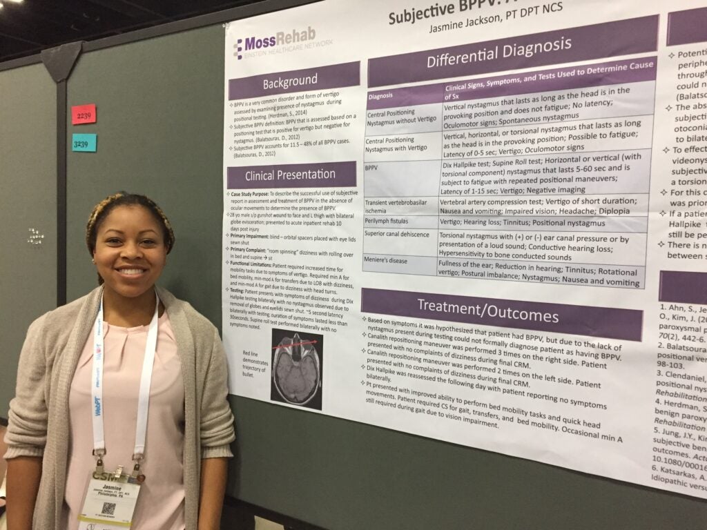 A woman with short dark and blonde braided hair wearing a tan cardigan over a light pink blouse standing in front of a poster.
