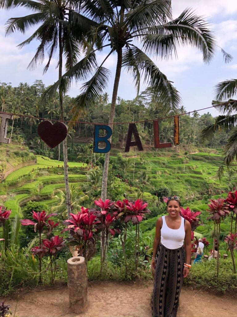 A woman with short dark hair wearing a white tank top and a black and tan patterned skirt standing in front of a garden in Bali.