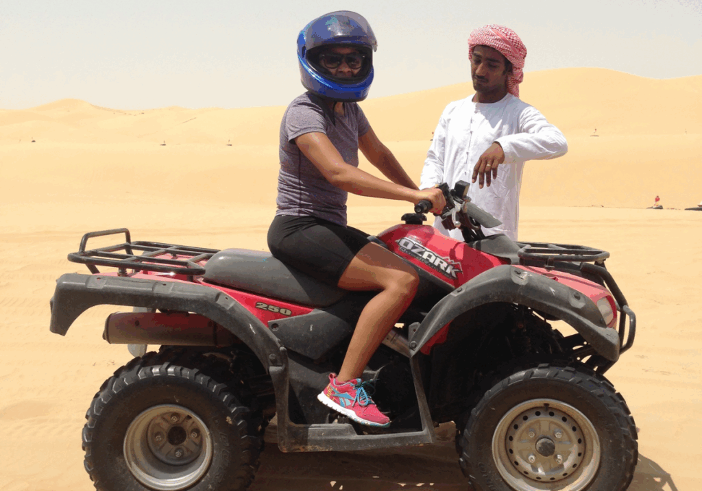 A woman wearing a blue helmet, a purple shirt and black shorts sitting on a red ATV while a man wearing a red head scarf and a white shirt looks on.