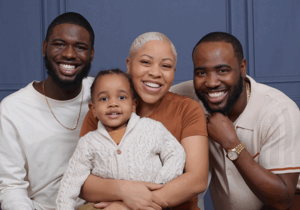 A man with short dark hair and a beard wearing a white shirt and a gold necklace, a woman with very short blonde hair wearing a brown shirt and holding a toddler wearing a cream sweater and a man with short dark hair wearing a tan shirt and a gold watch sitting together in front of a blue wall.