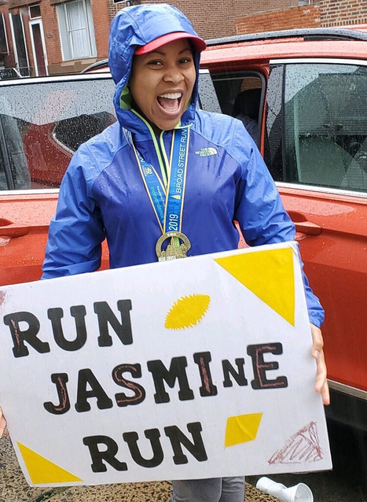 A woman wearing a blue rain jacket, gray pants and black shoes holding a sign that says "Run Jasmine Run"