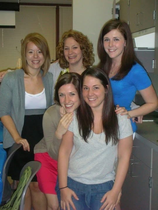A group of five women standing closely together next to chairs and cabinets.