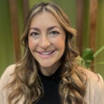 A woman with brown and blonde curled hair wearing a tan blazer over a black top.