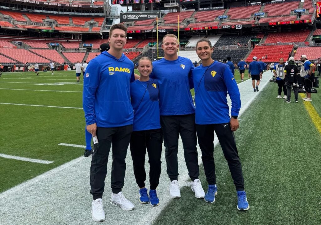A man with short brown hair, a woman with brown hair pulled back, a man with short light hair and a man with brown hair pulled back all wearing black pants and blue sweatshirts that have the Los Angeles Rams logo on them.