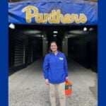 A woman with dark hair pulled back wearing a blue zip-up jacket and khaki pants holding a drink carrier of Gatorade bottles and standing in front of a sign that reads "Panthers."