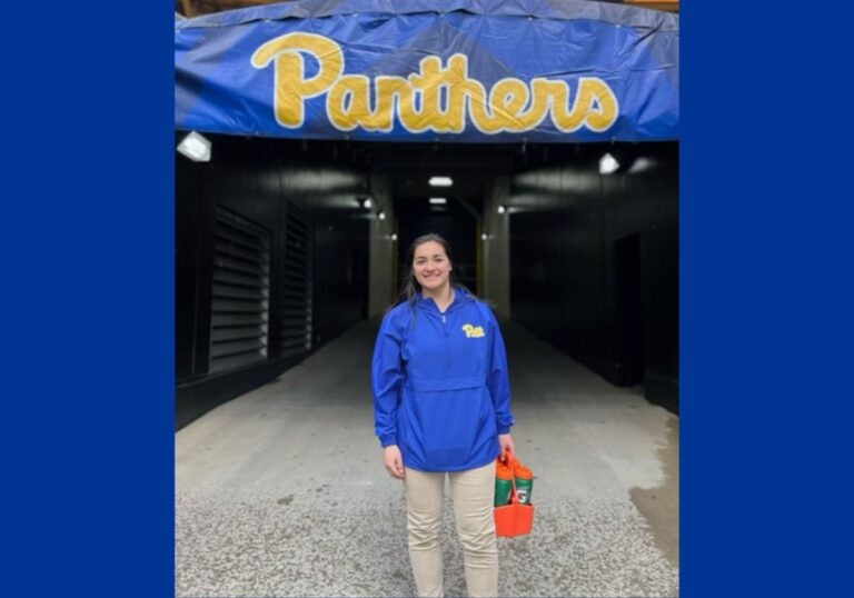 A woman with dark hair pulled back wearing a blue zip-up jacket and khaki pants holding a drink carrier of Gatorade bottles and standing in front of a sign that reads "Panthers."