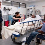 An older Black man lies on a hospital gurney in a hospital room as a man with a beard stands and two men sit in chairs looking at him