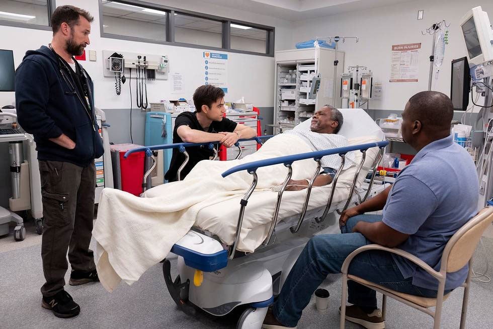 An older Black man lies on a hospital gurney in a hospital room as a man with a beard stands and two men sit in chairs looking at him
