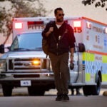 A man with a beard in a dark sweatshirt holding a backpack wearing sunglasses walks in front of an ambulance.