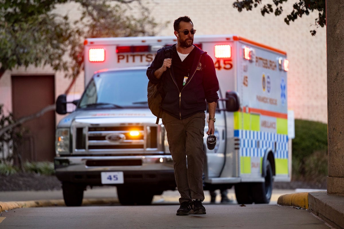 A man with a beard in a dark sweatshirt holding a backpack wearing sunglasses walks in front of an ambulance.